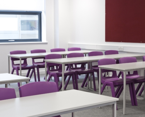 Empty classroom with purple chairs and white desks. There is a window at the back of the room.